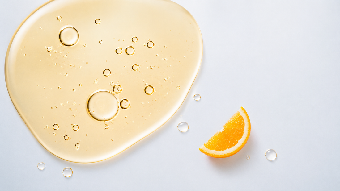 Minimal clinical wellness image with a golden liquid droplet and orange slice on a soft blue-gray background